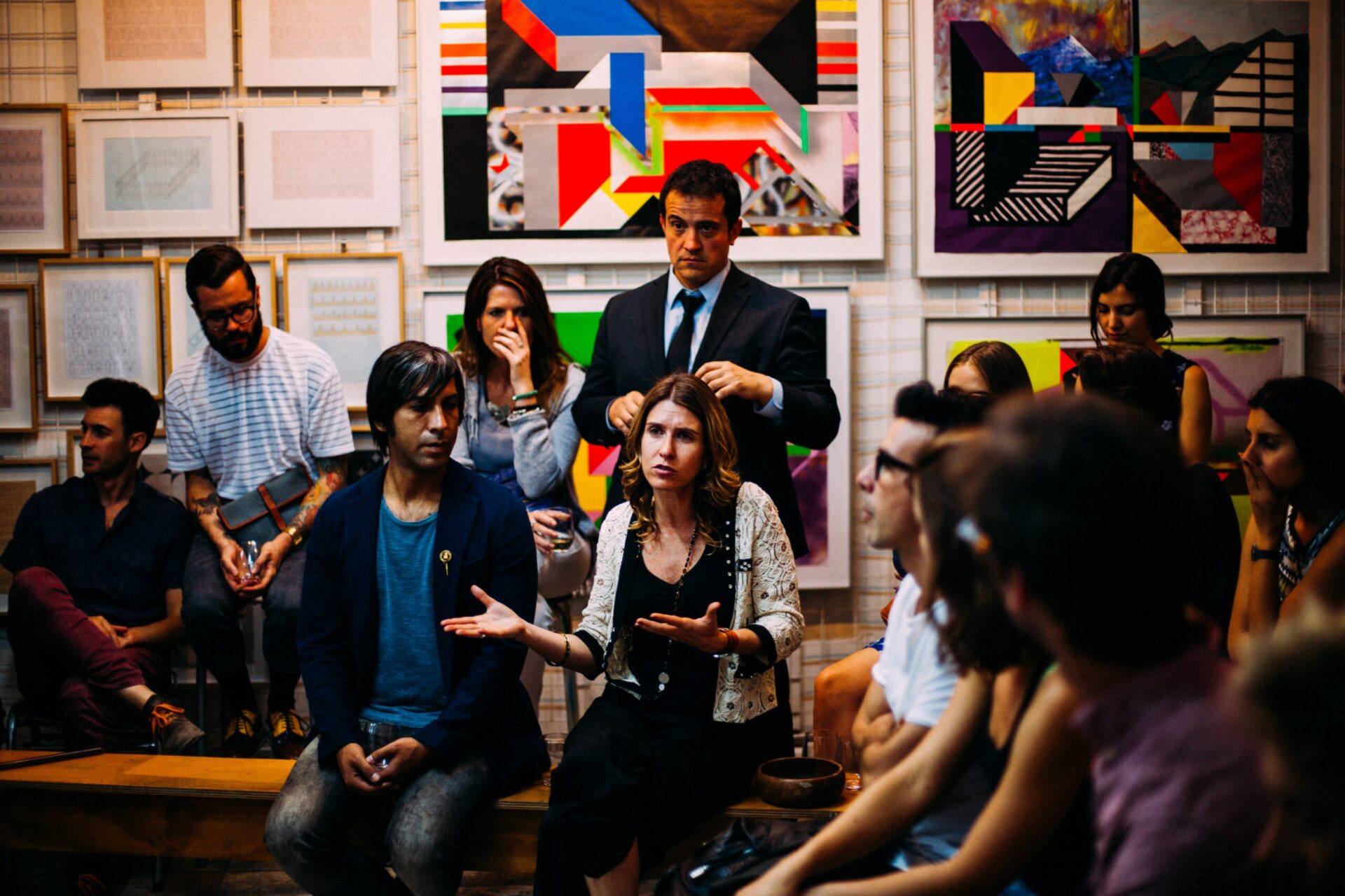 a diverse room full of people sit and stand in a semi-circle. One of the participants speaks and others look at her.